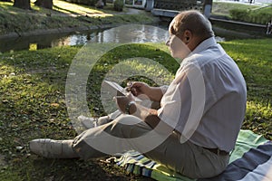 An elderly man in a white shirt is sitting on a blanket, on the ground in a park and reading an interesting book. A