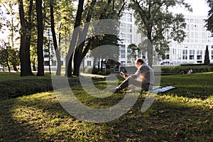 An elderly man in a white shirt is sitting on a blanket, on the ground in a park and reading an interesting book. A