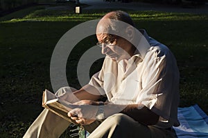 An elderly man in a white shirt is sitting on a blanket, on the ground in a park and reading an interesting book. A pensioner