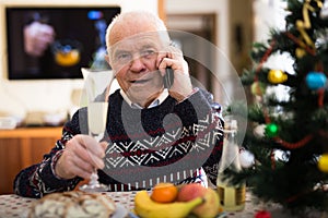 Elderly man wishes Merry Christmas by phone to friends and acquaintances sitting at the festive table in house