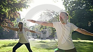 Elderly Man And Woman With Gray Hair Doing Fitness In Park. They Keep Their Arms Outstretched.
