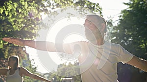 Elderly Man And Woman With Gray Hair Doing Fitness In Park. They Keep Their Arms Outstretched.