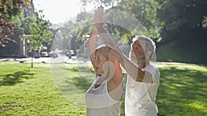 Elderly Man And Woman With Gray Hair Doing Fitness In Park. Man Corrects Woman Hands.