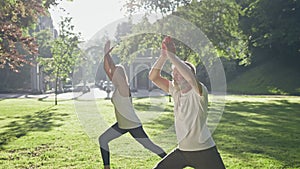 Elderly Man And Woman With Gray Hair Doing Fitness In Park. They Stand With One Foot Forward Hold Their Hands Up