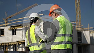 Elderly man and a young woman at a construction site. Builders in helmets and signal vests are discussing something.