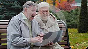 Elderly married couple sit on bench in autumn park make video call using laptop waving hands talking with grandchildren