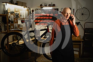 Elderly Mechanic in bicycle workshop using a mobile phone standing near bike