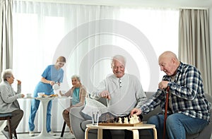 Elderly men playing chess while nurse serving breakfast to women. Assisting senior people