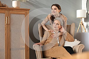 Elderly middle mother sitting in a chair and her daughter are hugging, looking away, trusted relations