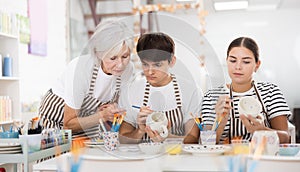 Elderly mother guiding young adult children painting pottery in family workshop