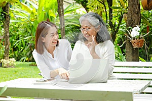 An elderly mother and her smiling, happy daughter sit and watch social media on a laptop computer in the garden.