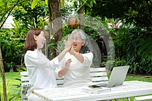 An elderly mother and her smiling, happy daughter sit and watch social media on a laptop computer in the garden.