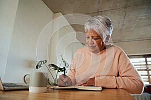 Elderly multi-ethnic female content while writing in her notebook with her pen in front of laptop. Sitting at counter at