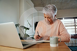 Elderly multi-ethnic female writing in notebook with pen while sitting in front of laptop at modern counter.