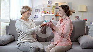 Elderly mum giving present to daughter sitting sofa at home, relations closeness