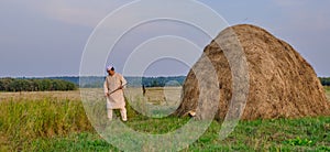 An elderly Muslim man in an embroidered skullcap and white traditional Clothes mows hand-scythe grass in a hayfield