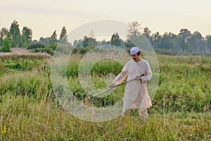 An elderly Muslim man in an embroidered skullcap and white traditional Clothes mows hand-scythe grass in a hayfield