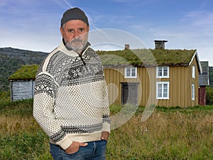 A Norwegian with a sweater is standing in a meadow in front of a typical old wooden house with a grass roof.