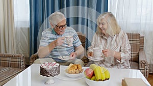 Elderly old couple holding drinking tea talking, laughing, smiling, kissing, sitting on sofa at home