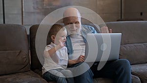 Elderly old man together with his grandson boy sit together on sofa side of laptops, waving greetings with their hands