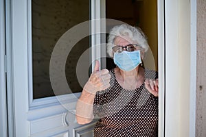 Elderly old senior woman opening front door of house and welcoming people at home wearing a surgical mask to avoid covid19 and cor