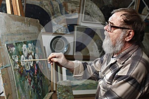 Elderly painter with beard and glasses draws a flowers picture by oil paint in art workshop