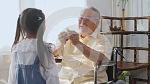 Elderly person sitting lonely in a wheelchair, sick and granddaughter running come holding a paper bird to play