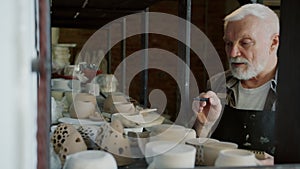 Elderly potter in dirty apron counting pots in workshop writing information in notebook