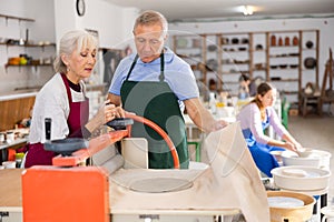 Elderly potter teaches woman how to roll clay on a craft machine clay press roller