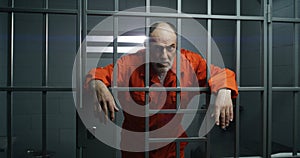 Elderly prisoner in orange uniform holds hands on metal bars, looks at camera