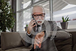 Elderly professional checking time while talking on smart phone while waiting for a train. Commuting to work