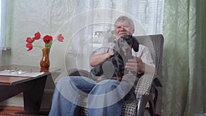 Elderly retired man with gray mustache is sitting in cozy rocking chair and stroking cute dachshund puppy on his lap in