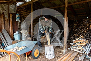 An elderly Russian man hunched wearily chopping wood with an axe in a barn