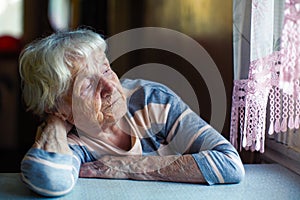 An elderly sad woman sits sadly near the window.