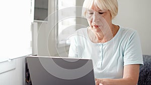 Elderly senior blond woman working on laptop computer at home. Remote freelance work on retirement