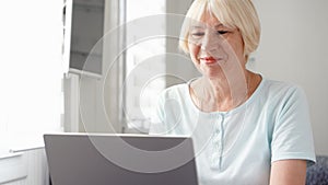 Elderly senior blond woman working on laptop computer at home. Remote freelance work on retirement