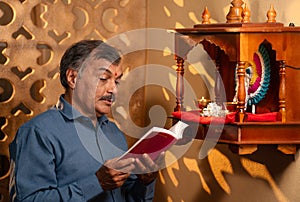 elderly senior man reading hymns from religious book in front of god near mandapam at home at morning - concept of