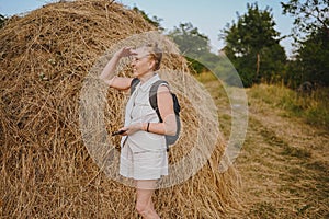 Elderly senior traveling backpacker mature woman tourist smiling posing with stack of hay in field outdoors in