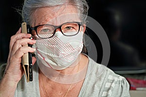 Elderly senior woman with glasses wearing hand made cotton mouth nose virus face mask, talking over her phone. Coronavirus covid-