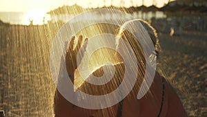 Elderly senior woman with gray hair taking morning shower at the beach, washing her face, sun backlight shines in background