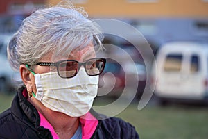 Elderly senior woman wearing home made cloth face mouth nose virus mask outside, blurred buildings and cars background. Can be