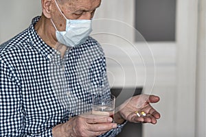 Elderly sick man in a medical mask. He holds pills and a glass of water in his hands.