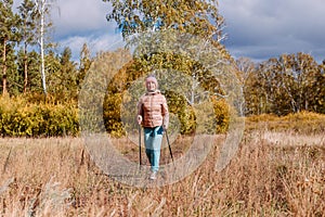 Elderly sixty-year-old woman in sportswear is engaged in Nordic walking with sticks in the forest in the autumn