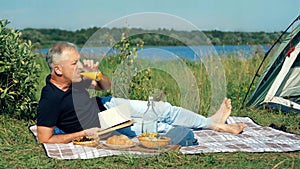 An elderly slender man is sitting on the grass by the pond, reading a book and drinking orange juice on a sunny summer