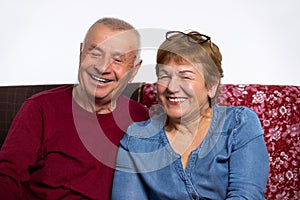 An elderly smiling married couple looking at the camera, sitting home on couch