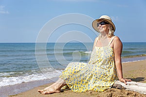 An elderly smiling woman in a yellow sundress, sunglasses and a hat sits on the seashore. Active lifestyle, travel and relaxation