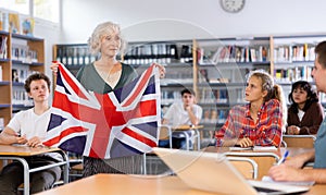 Elderly teacher shows the flag of Great Britain to teenagers and talks about this country during lesson at library