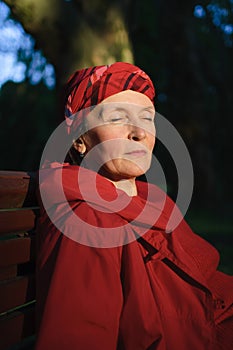Elderly tired woman in red clothes in park sitting on the bench and enjoying a good weather and sunset in the park