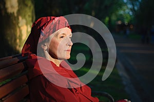 Elderly tired woman in red clothes in park sitting on the bench and enjoying a good weather and sunset in the park