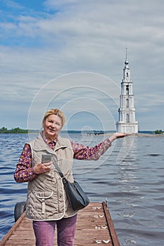 An elderly tourist poses against the background of the bell tower and pretends to hold the tower in the palm of her hand